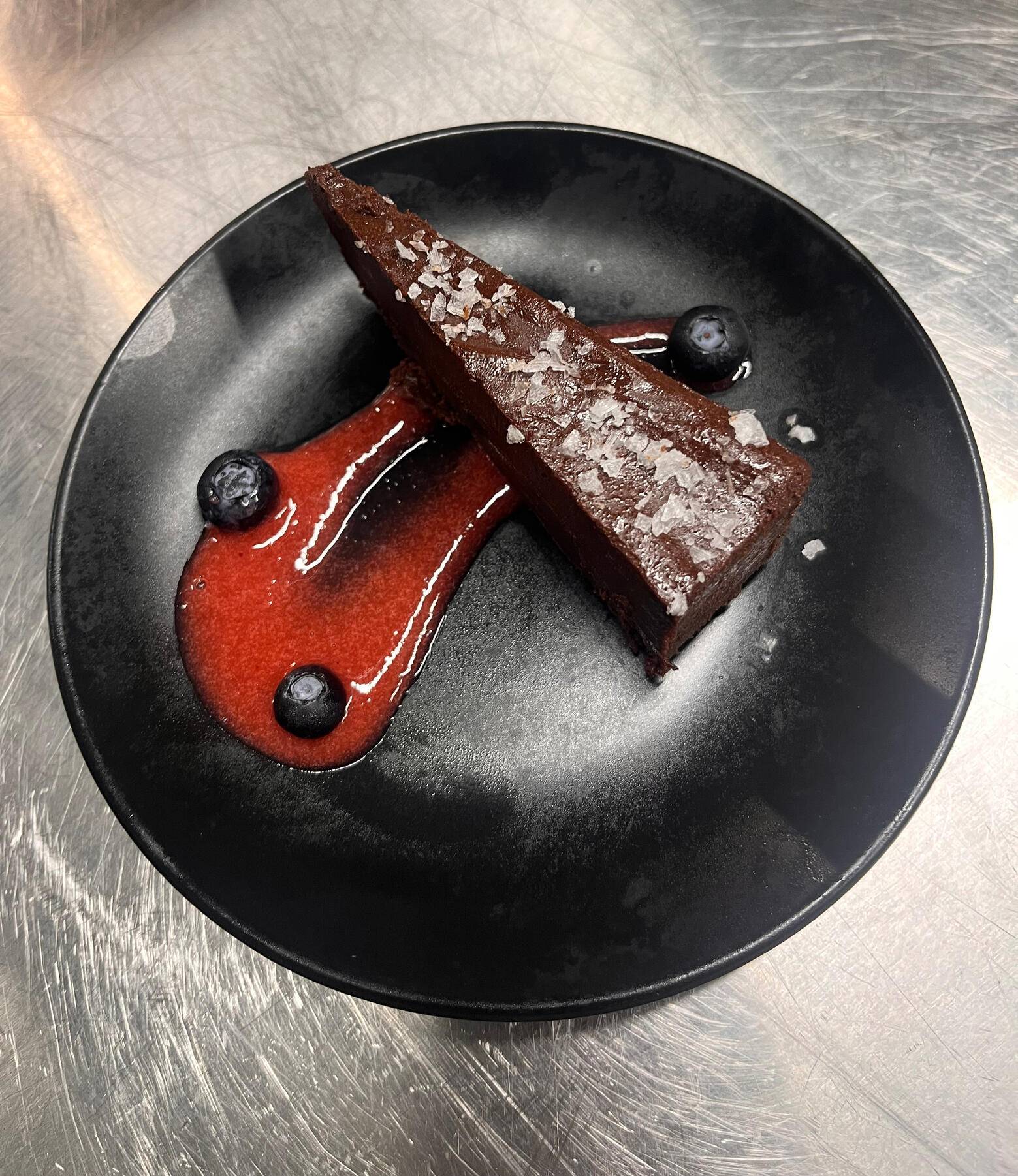 A slice of chocolate cake with flaky salt, strawberry sauce and blueberries on a black plate
